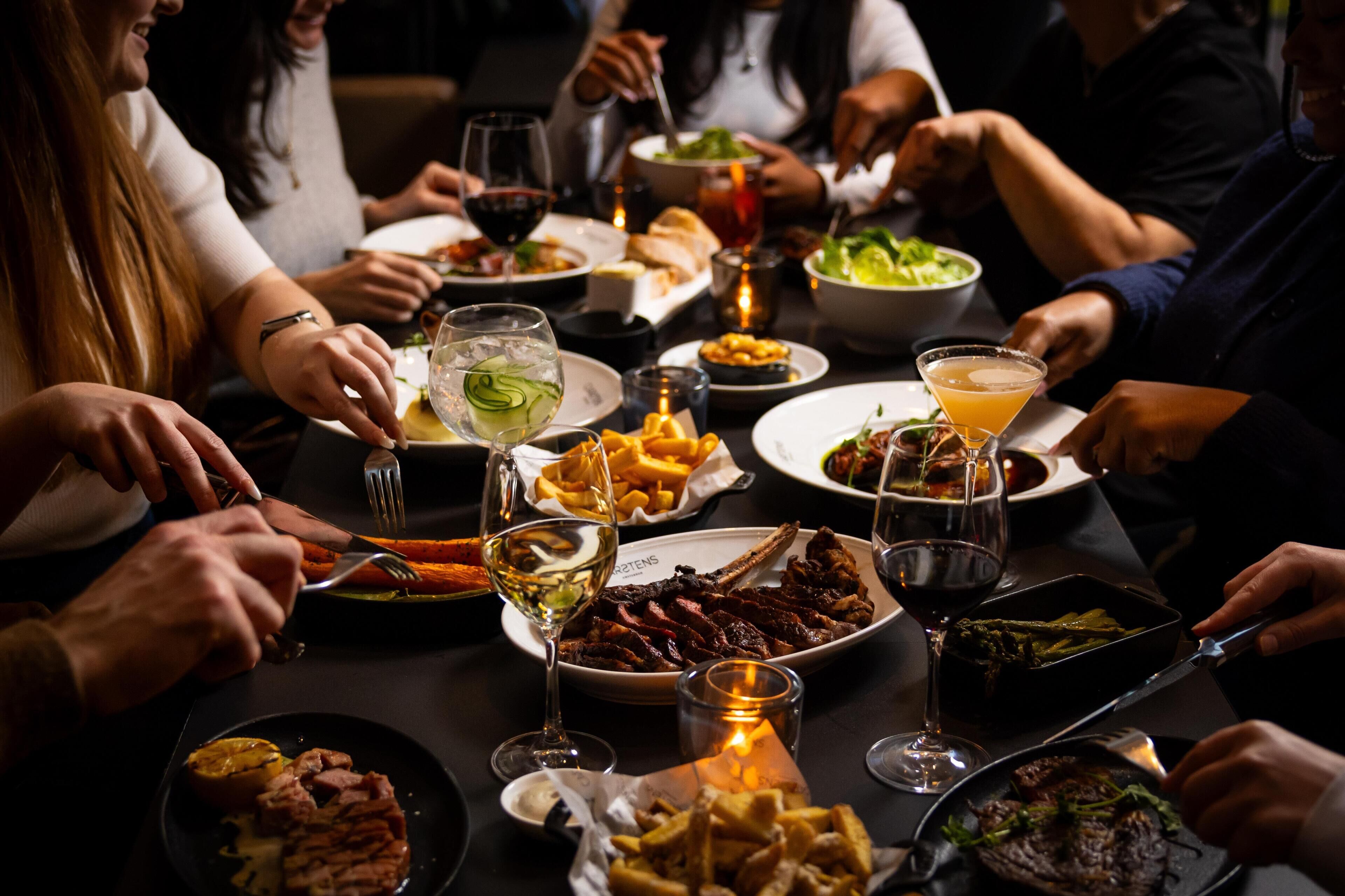 Un groupe de personnes est réuni autour d’une table, où l’on peut voir différents plats, des verres de vin et des bougies.