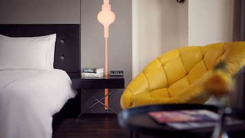 Habitación de hotel con cama, mesita de noche con libros y un reloj, un sillón amarillo y una lámpara moderna.