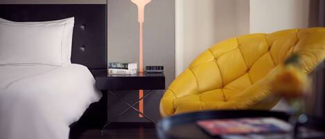 A hotel room with a bed, a bedside table with books and a clock, a yellow armchair, and a modern lamp.