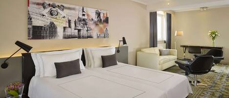 A hotel room with a large bed, a desk, a chair, and a couch. There is a painting on the wall and a vase with flowers on the desk.