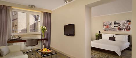 Habitación de hotel con cama, escritorio con silla, pequeña mesa con fruta, ventana con cortinas y televisor montado en la pared.
