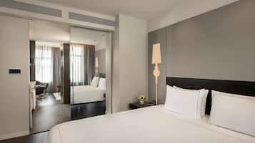 A modern hotel room with a large bed, a bedside table with a lamp, a mirror, and a view of the hallway and another room.
