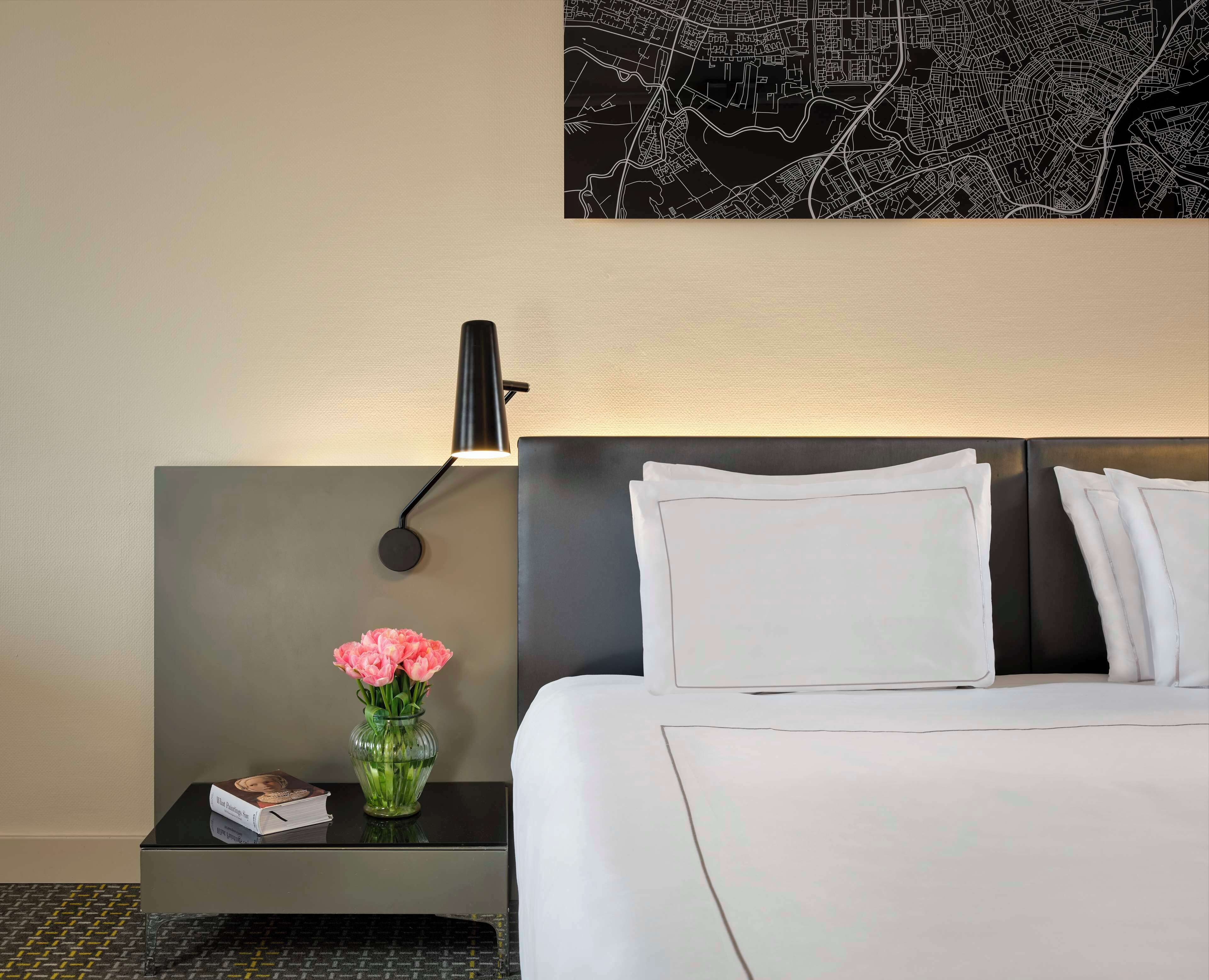 A hotel room with a neatly made bed, a bedside table with a book and a vase of flowers, a wall-mounted lamp, and a map on the wall.