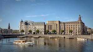 Un hotel frente al río con un gran edificio, una torre de reloj y un río con barcos.