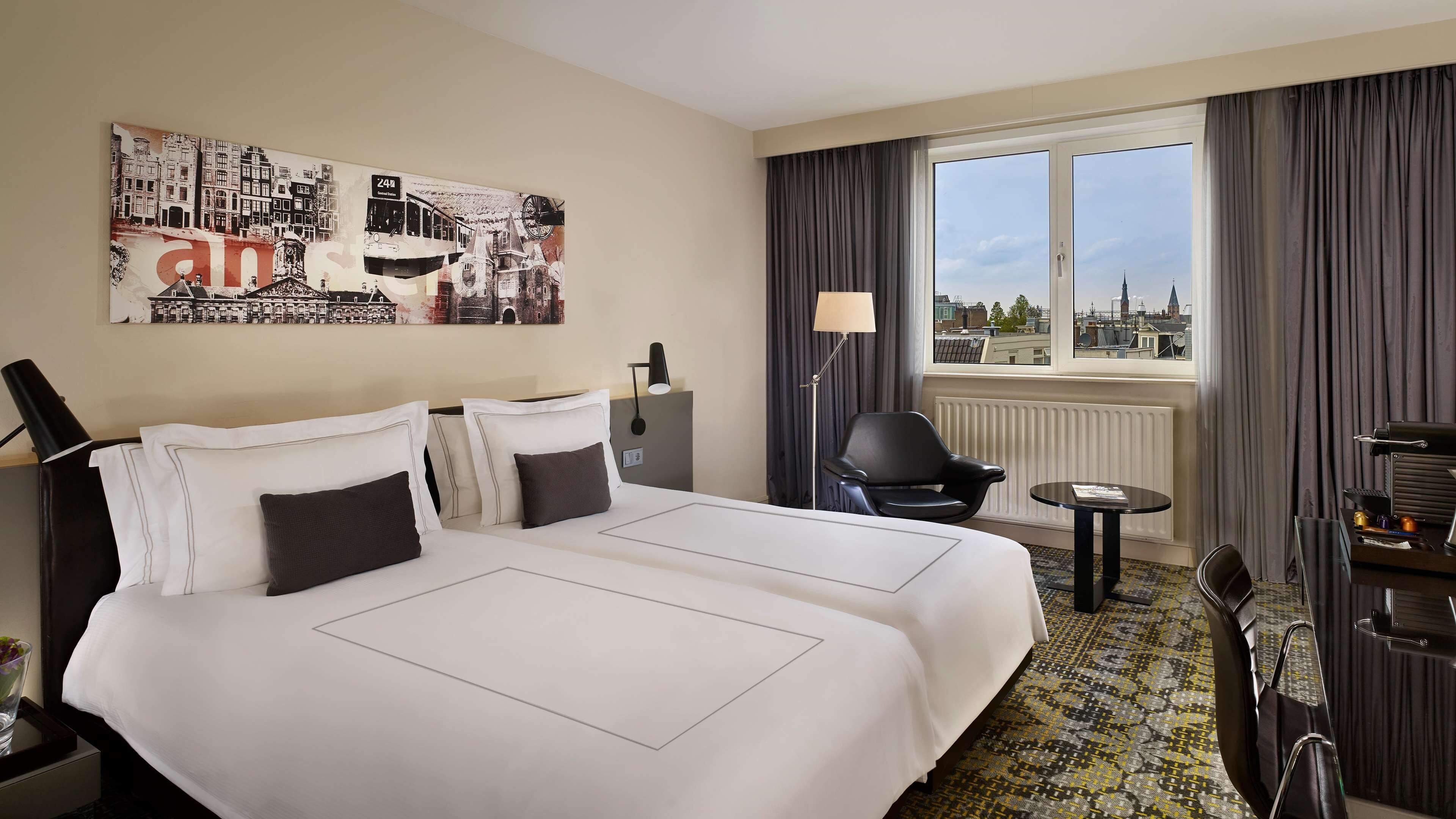 A hotel room with two beds, a desk, a chair, a lamp, and a view of the city.