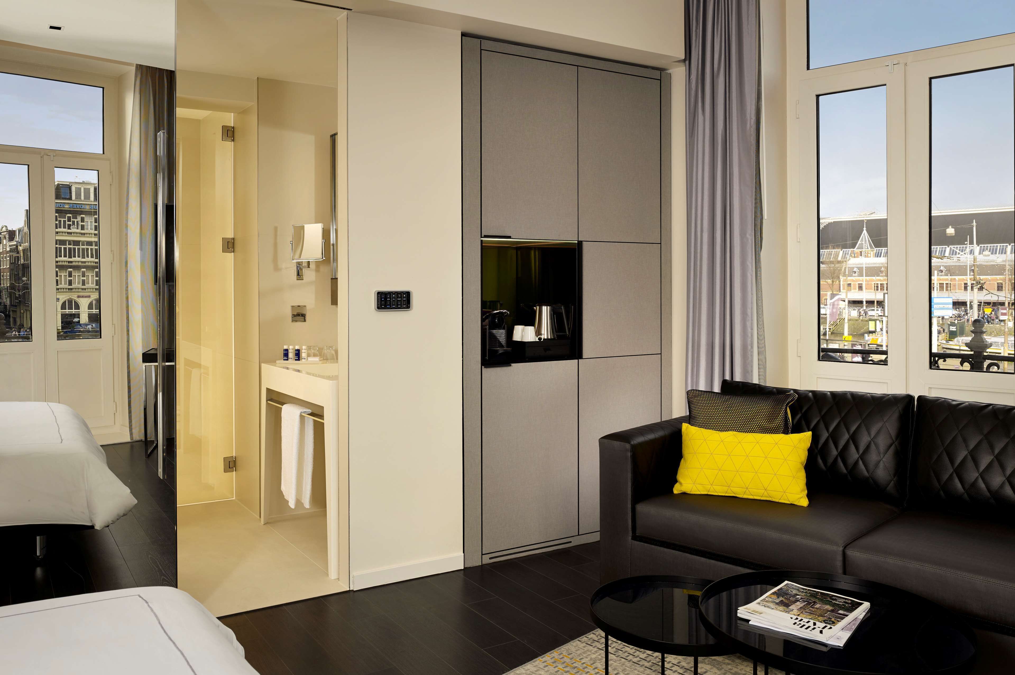 A modern hotel room with a sofa, a small table, a bathroom with a mirror, and a view of the city.