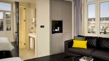 A modern hotel room with a sofa, a small table, a bathroom with a mirror, and a view of the city.