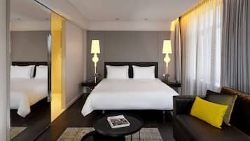 A modern hotel room with a large bed, two bedside tables with lamps, a sofa with a yellow pillow, and a small coffee table.