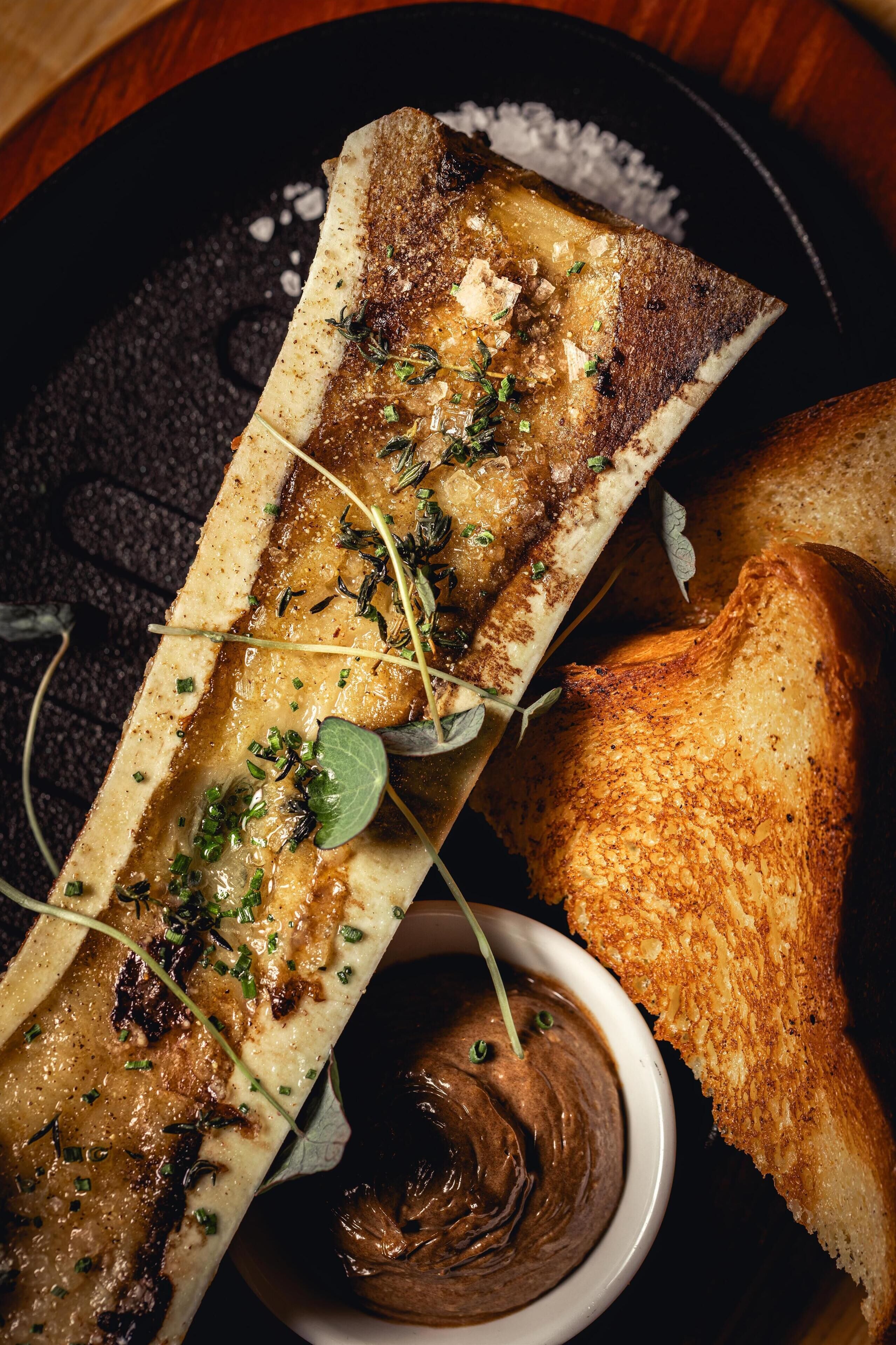 A dish with a bone, bread, and a small bowl of sauce.