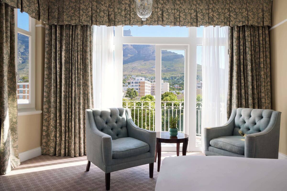 Habitación de hotel con una cama grande, dos sillones, una mesita y un balcón con vistas a edificios y montañas.