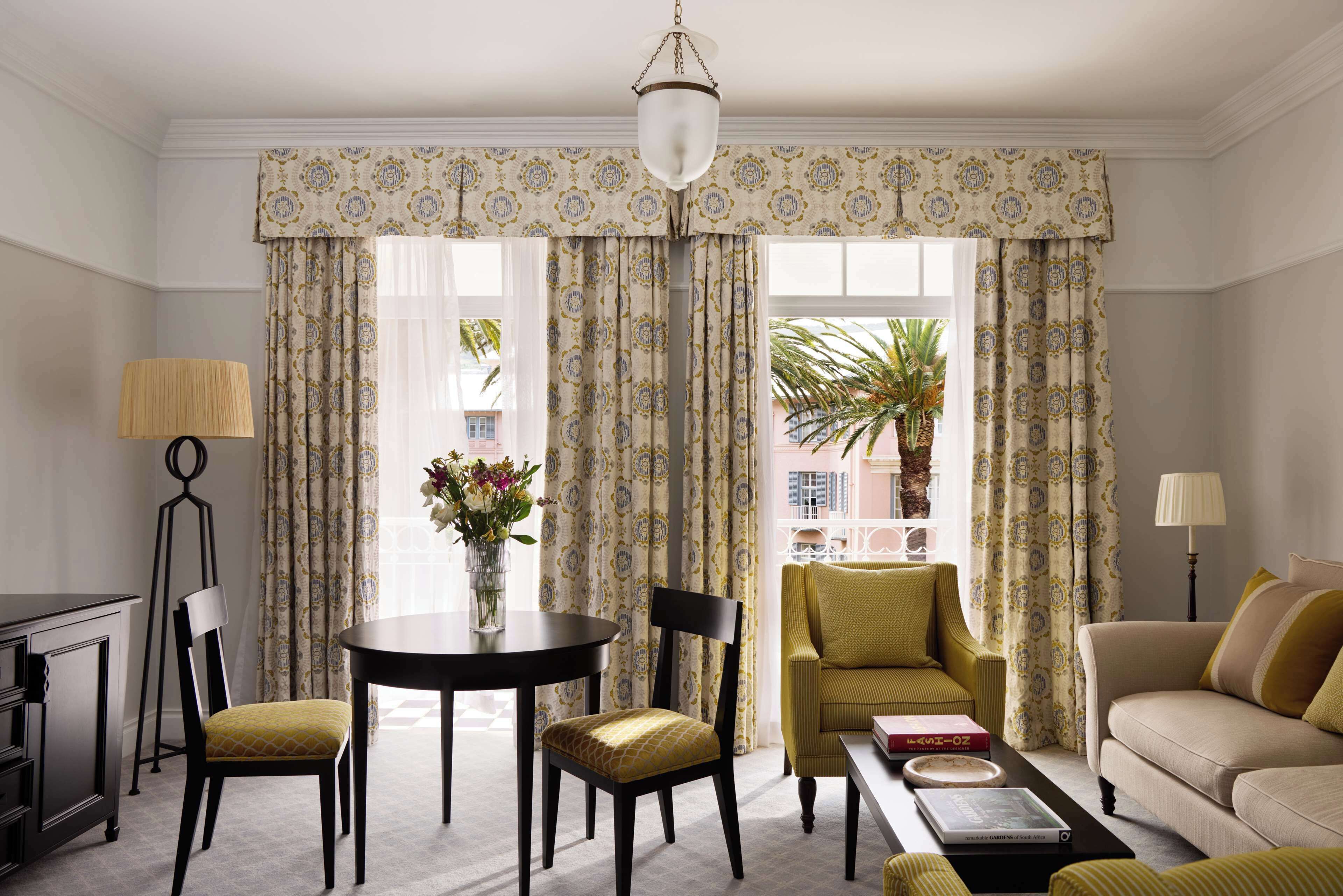 A well-lit room with a dining table, two chairs, a sofa, and a coffee table. A lamp is on the side table, and there is a window with curtains.