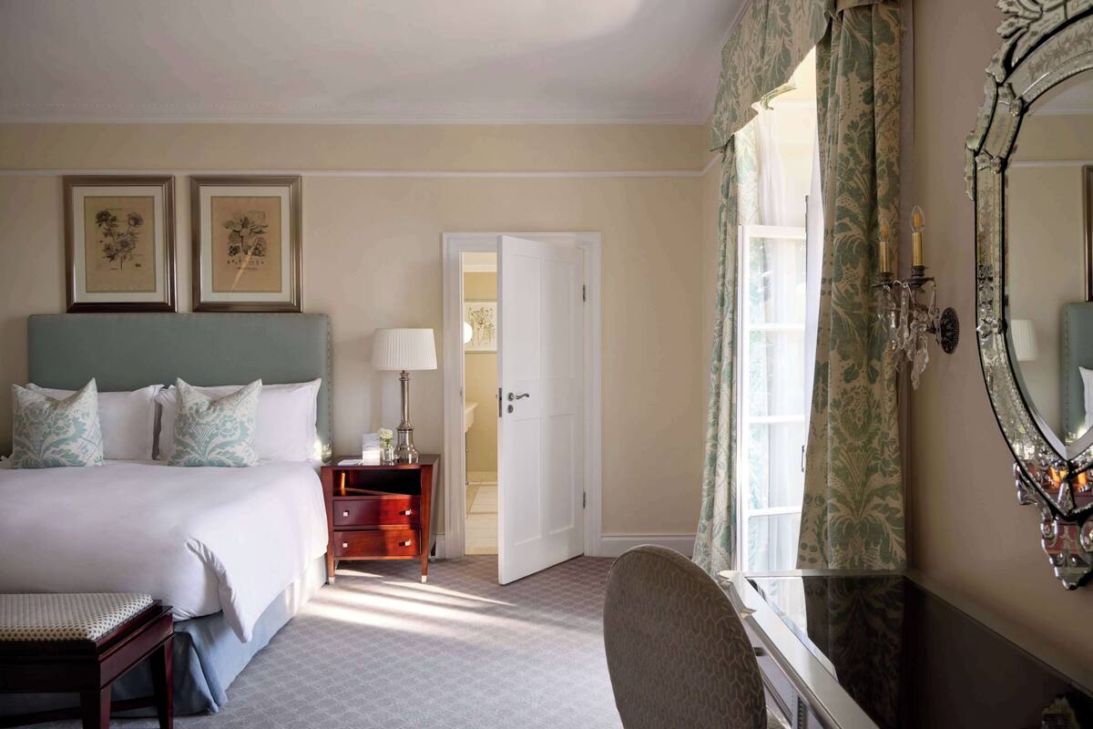 Una habitación de hotel con cama, mesita de noche, silla, cómoda, espejo y puerta que da a otra estancia.