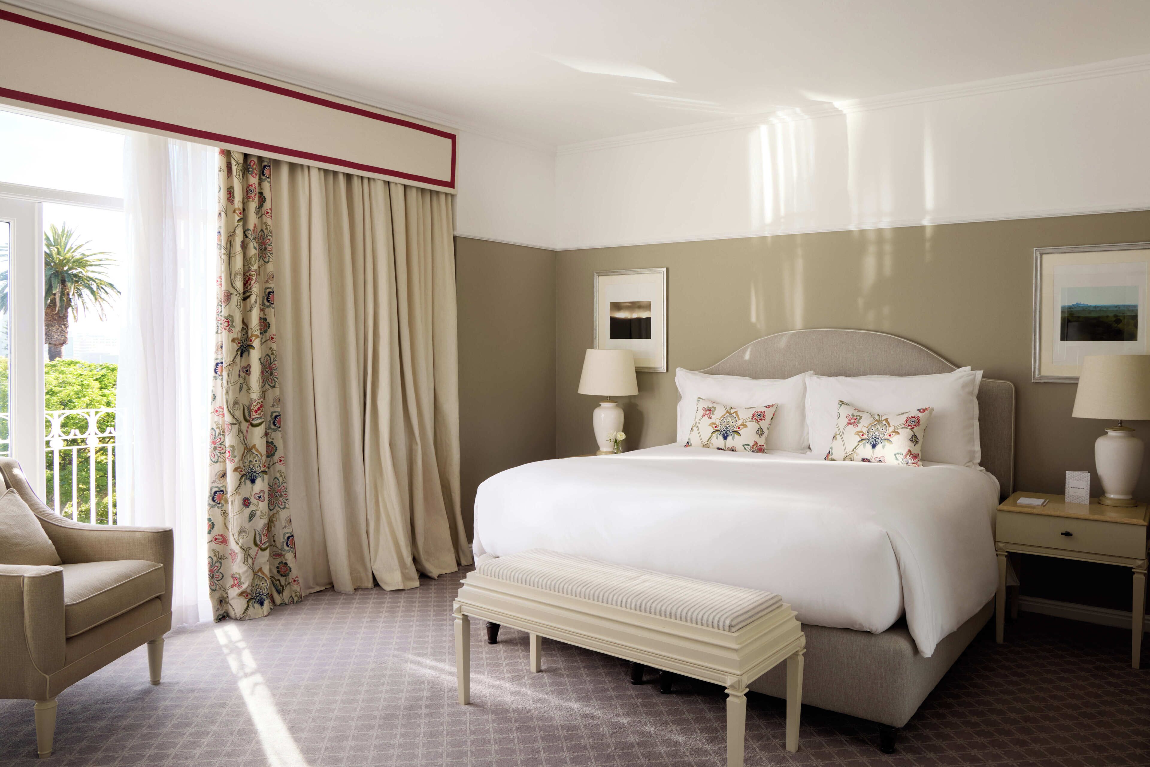 Habitación de hotel con una cama grande, un banco, una silla, dos mesitas de noche con lámparas y un ventanal con cortinas.