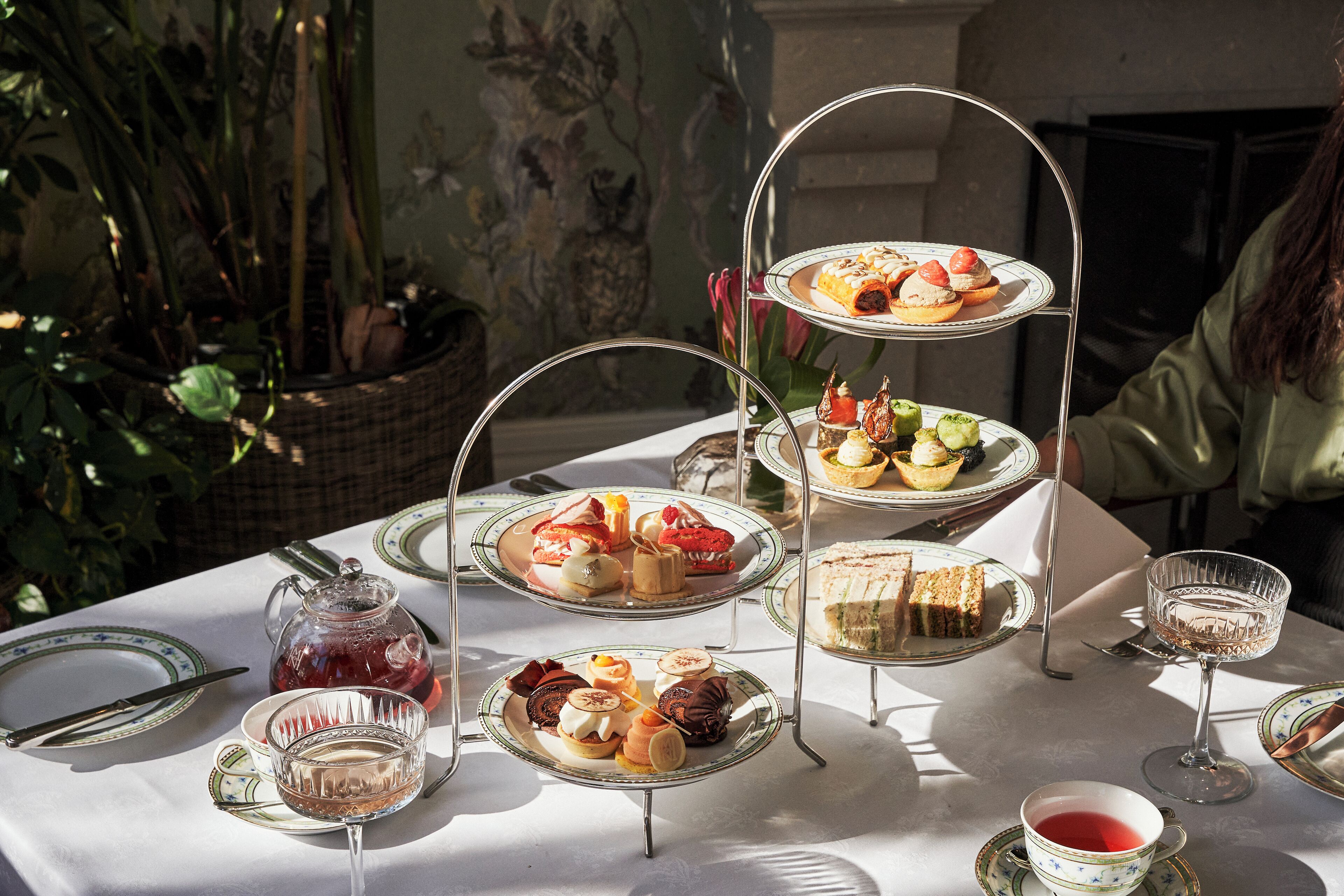 A three-tiered dessert stand with various pastries and sandwiches, a pitcher of red beverage, and a glass of white beverage on a table with a white tablecloth.