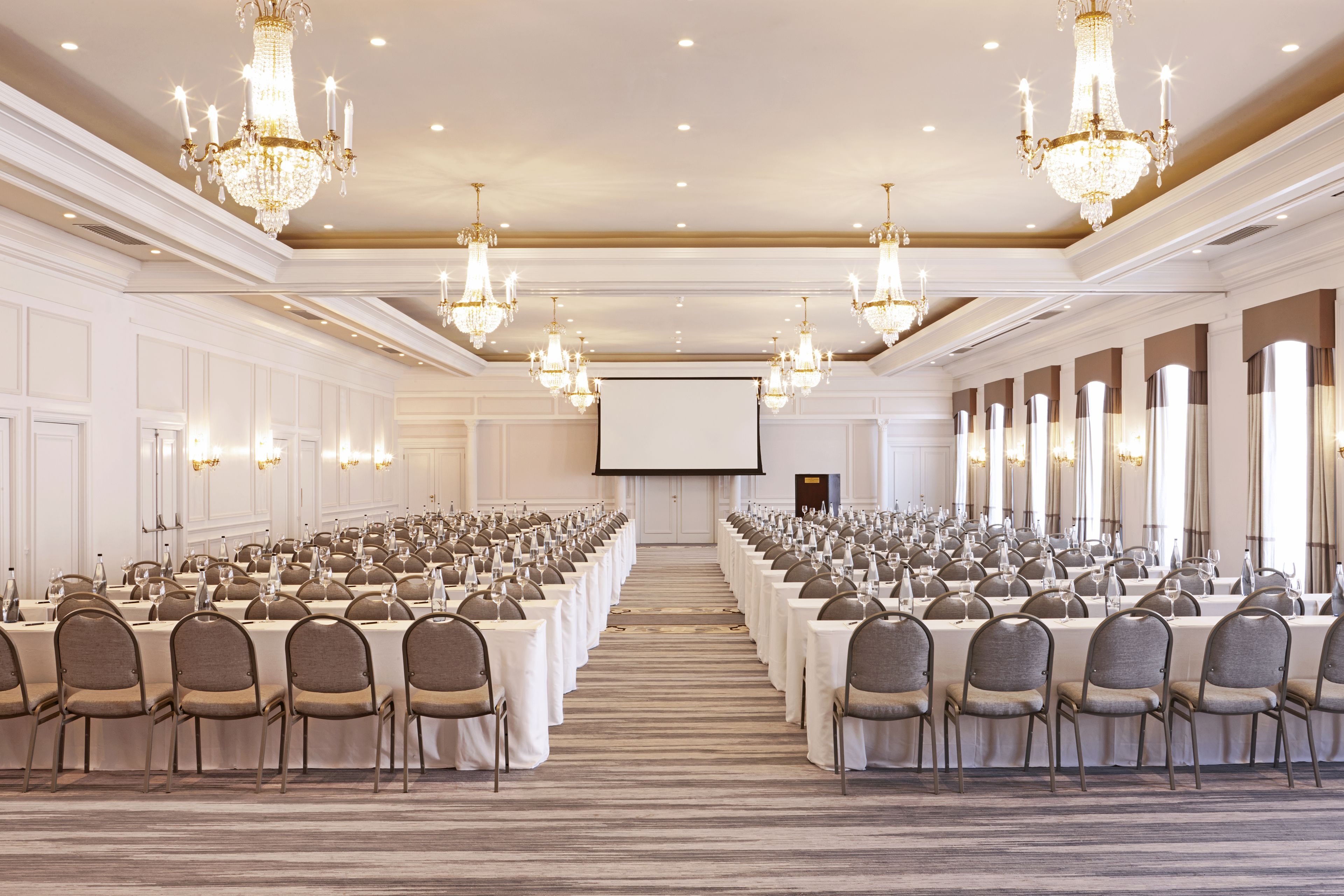 Een ruime conferentiezaal met rijen tafels en stoelen, kroonluchters en een scherm voor een beamer.