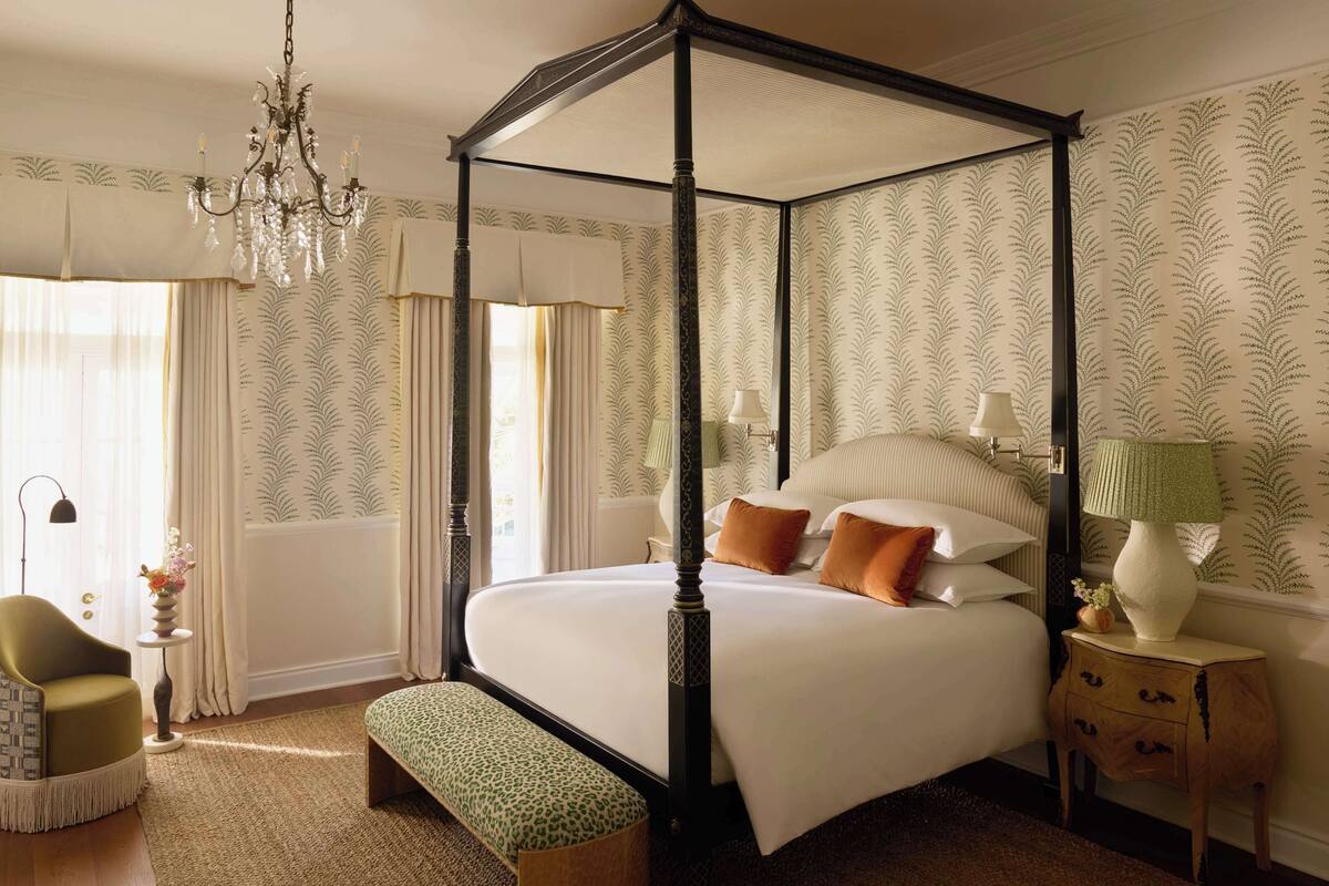 Una cama con dosel y cortinas, un candelabro, una mesita de noche con lámpara y un banco.