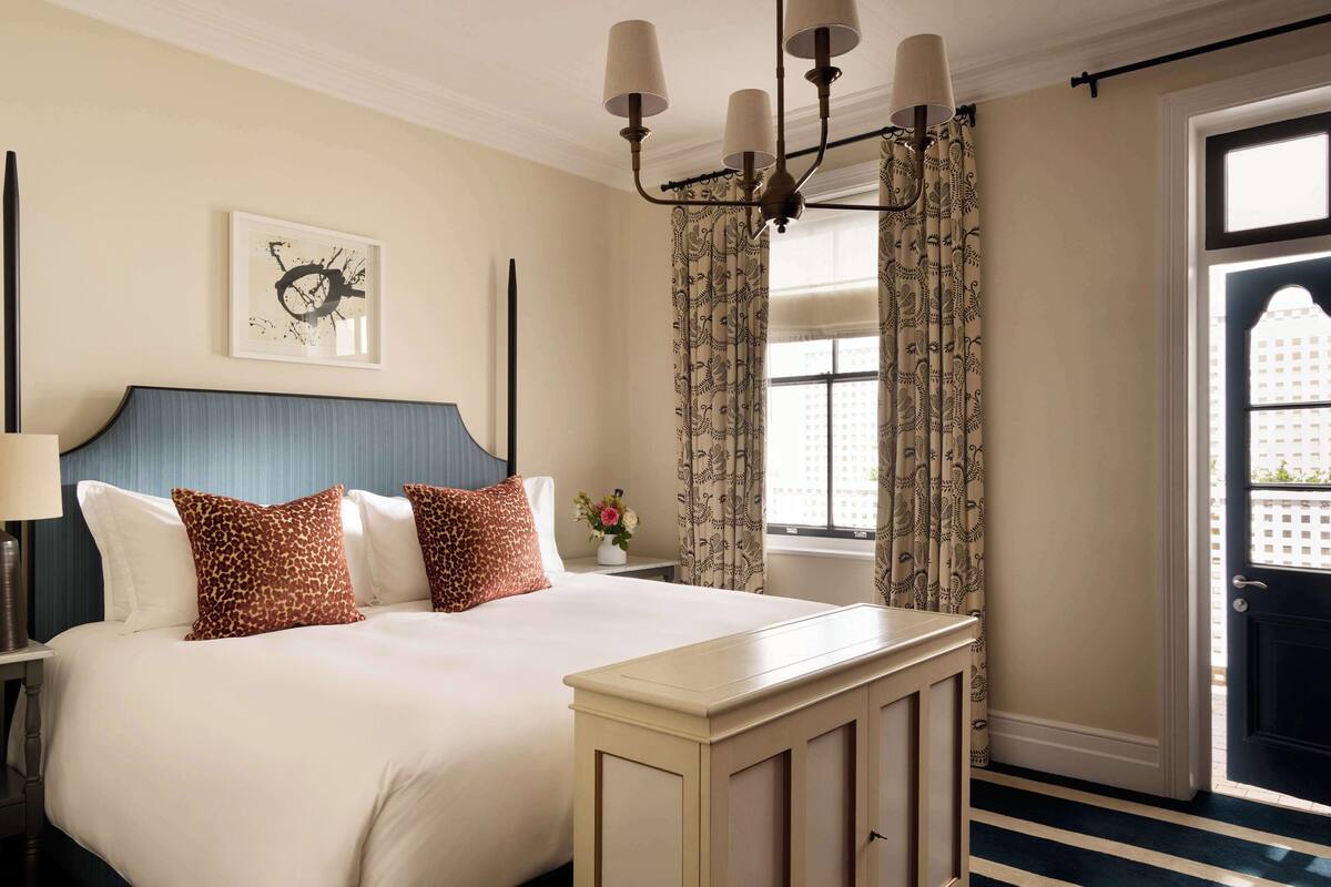 Una habitación de hotel con una cama grande, una mesita de noche con lámpara, un reloj de pared y una ventana con cortinas estampadas.