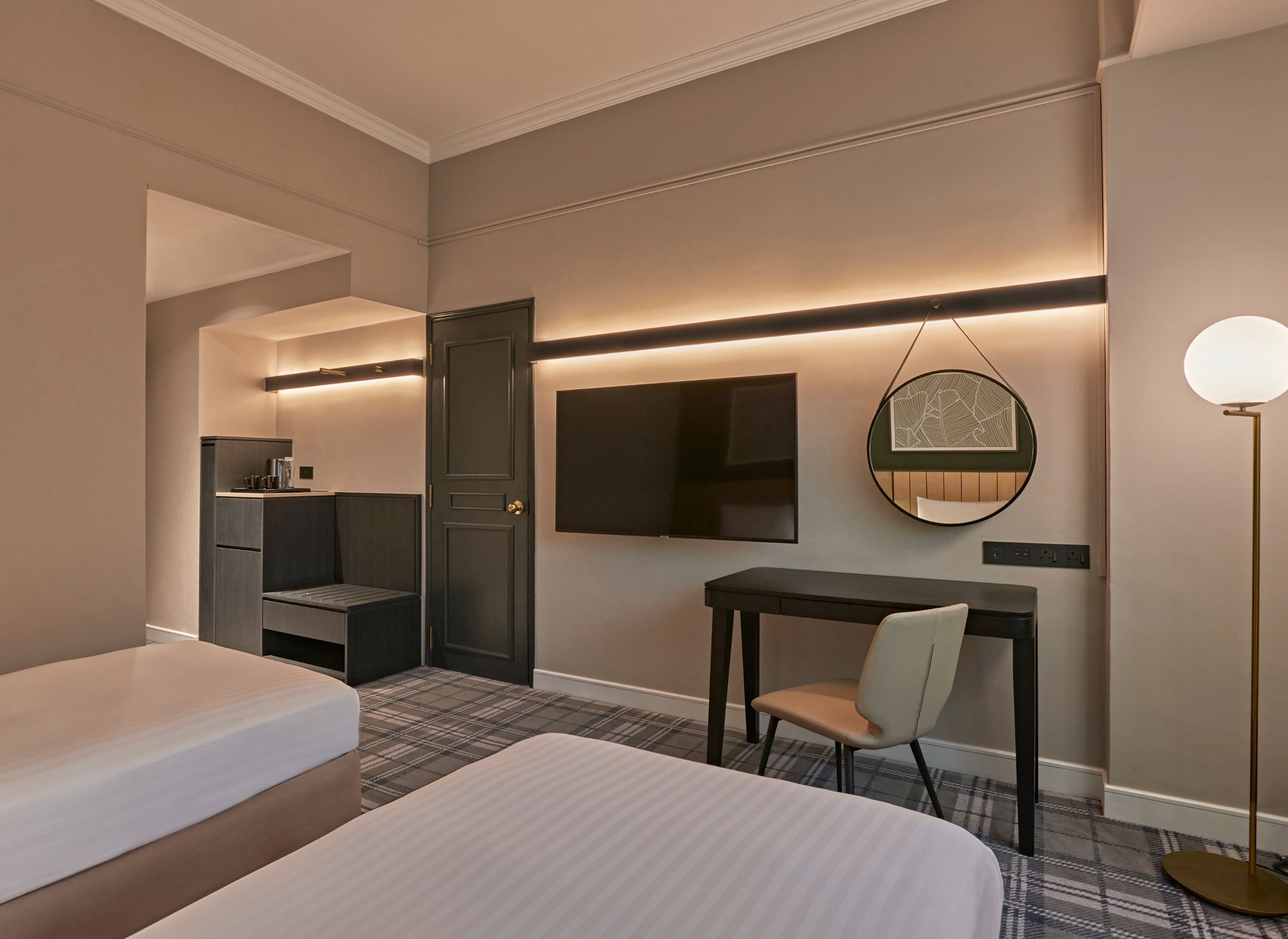 Habitación de hotel con cama, escritorio, silla, armario, espejo y lámpara de pie.