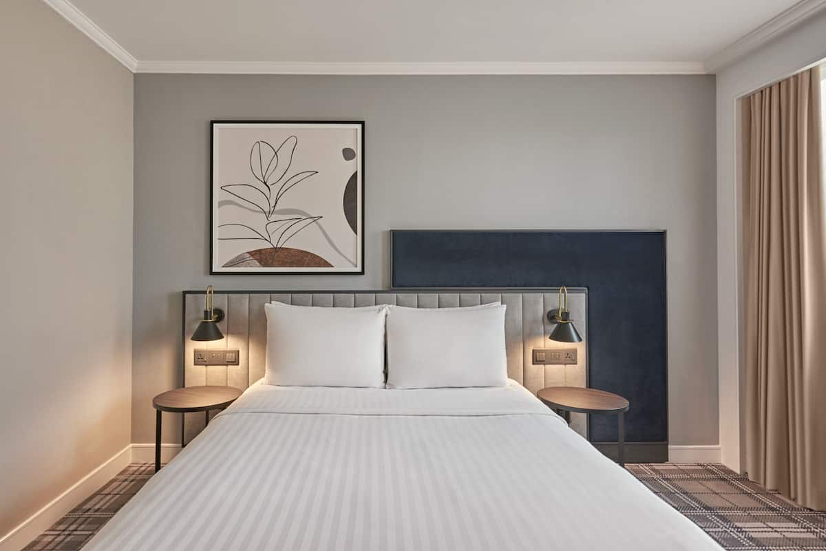 Una habitación de hotel con una cama grande, dos mesitas de noche con lámparas, un cabecero, una obra de arte enmarcada y una ventana con cortinas.