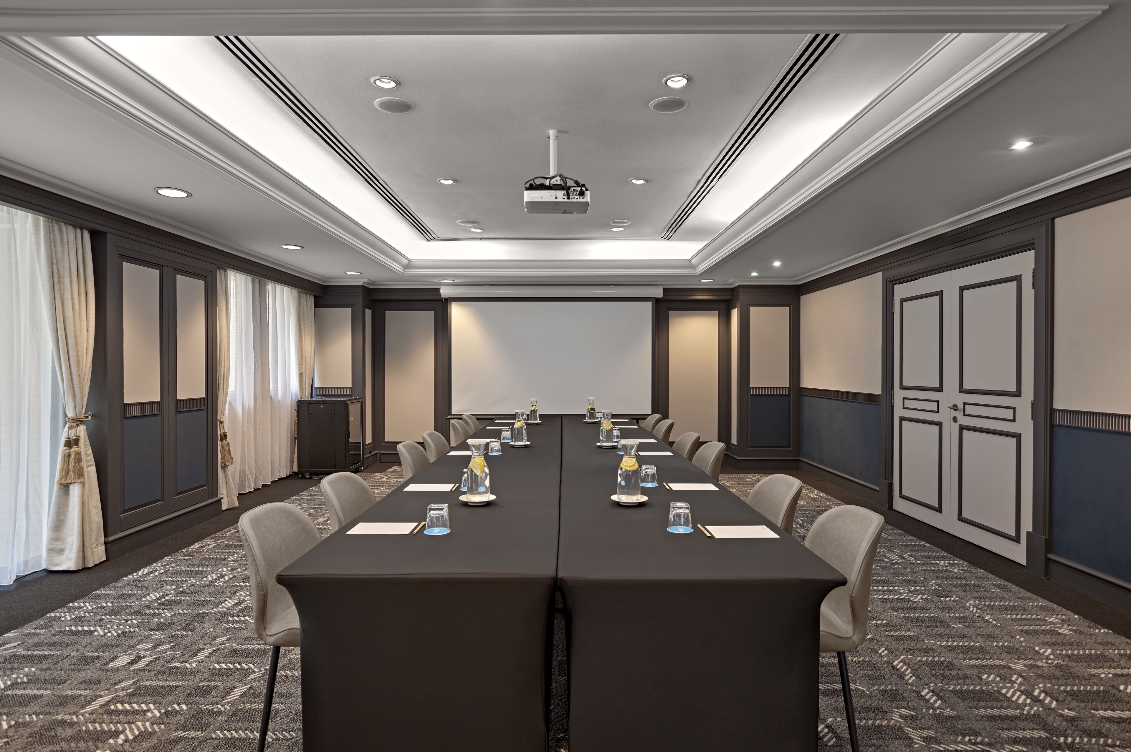 Un salón de conferencias con una larga mesa, sillas, un proyector y una pantalla.