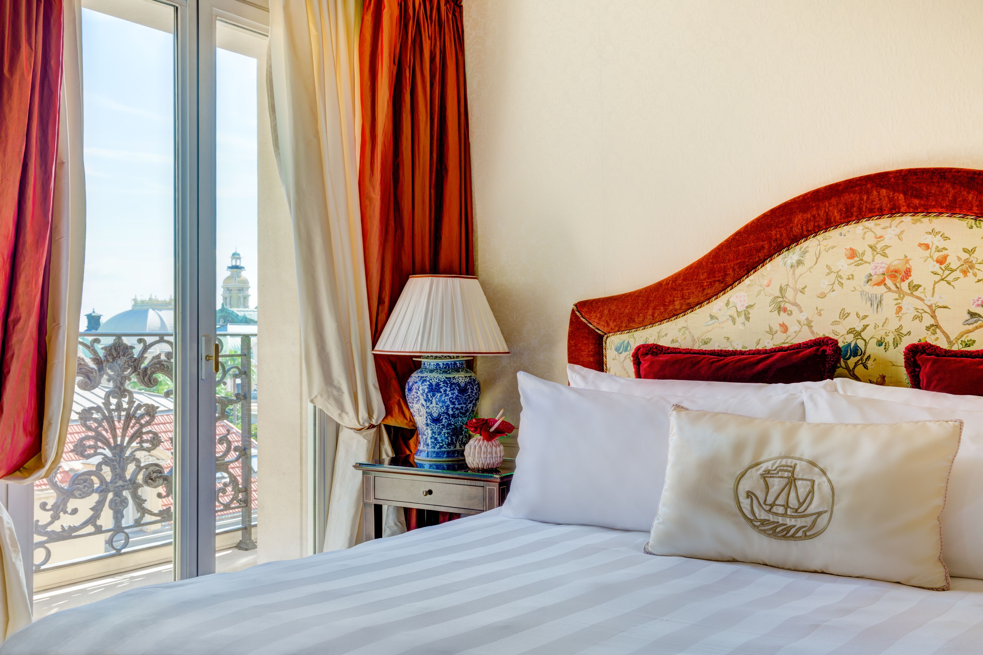 Ein Hotelzimmer mit einem Bett, einem Nachttisch, einer Lampe und Blick auf eine Stadtkulisse durch große Fenster.
