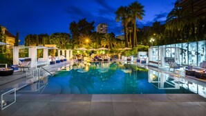 A pool area with lounge chairs and umbrellas, surrounded by palm trees and a building in the background. - Hôtel Métropole Monte Carlo – Spa Guerlain (Monaco)