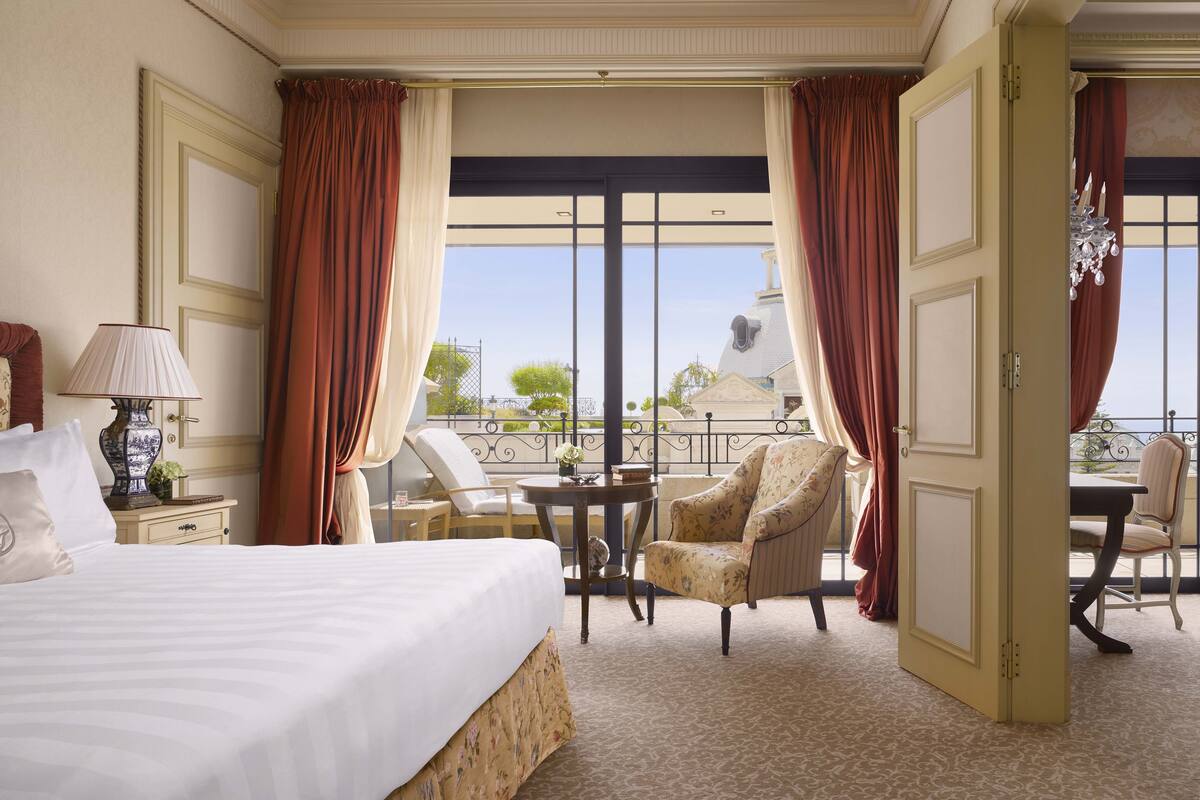 A hotel room with a large bed, a sitting area with a chair and a table, and a view of a building through the windows.
