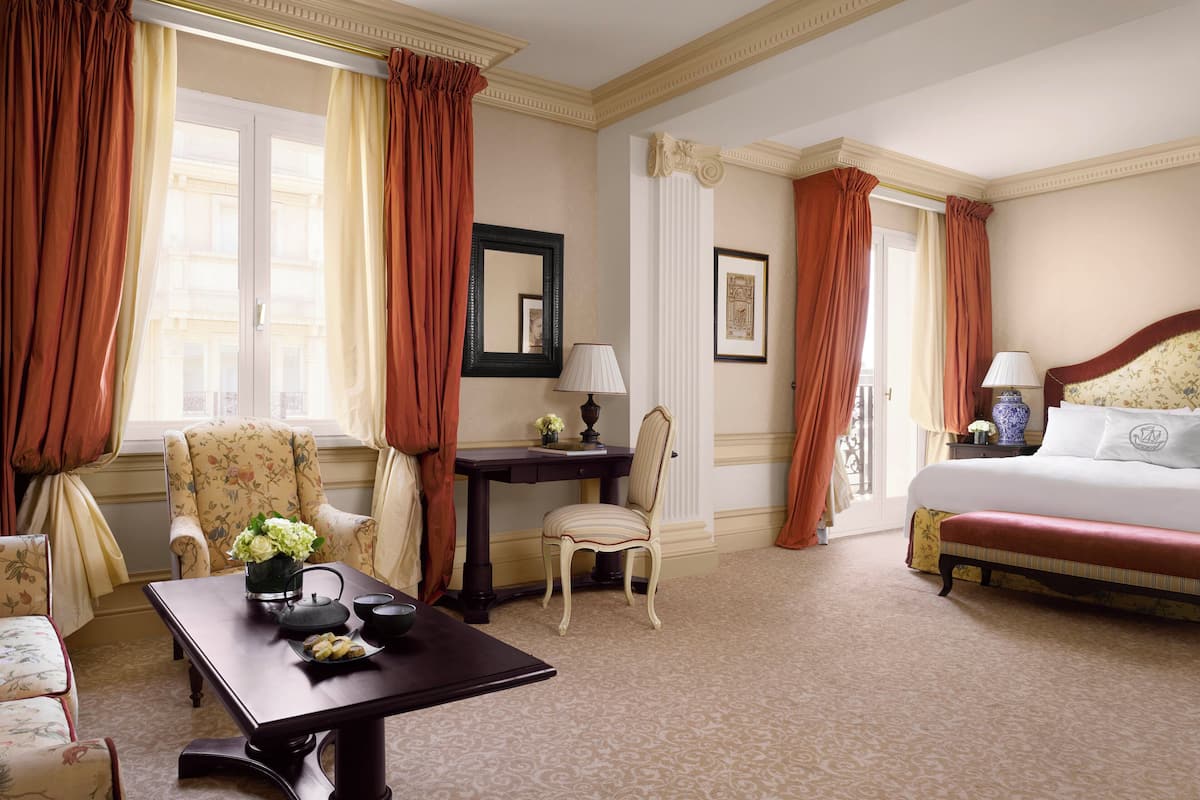 A hotel room with a bed, a desk, a chair, a table with a vase of flowers, and a window with curtains.