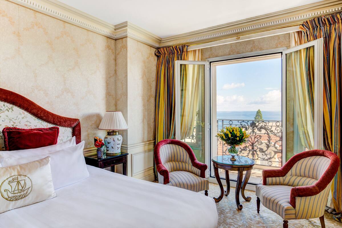 A hotel room with a large bed, two armchairs, a small table with a vase, and a view of the city through large windows.