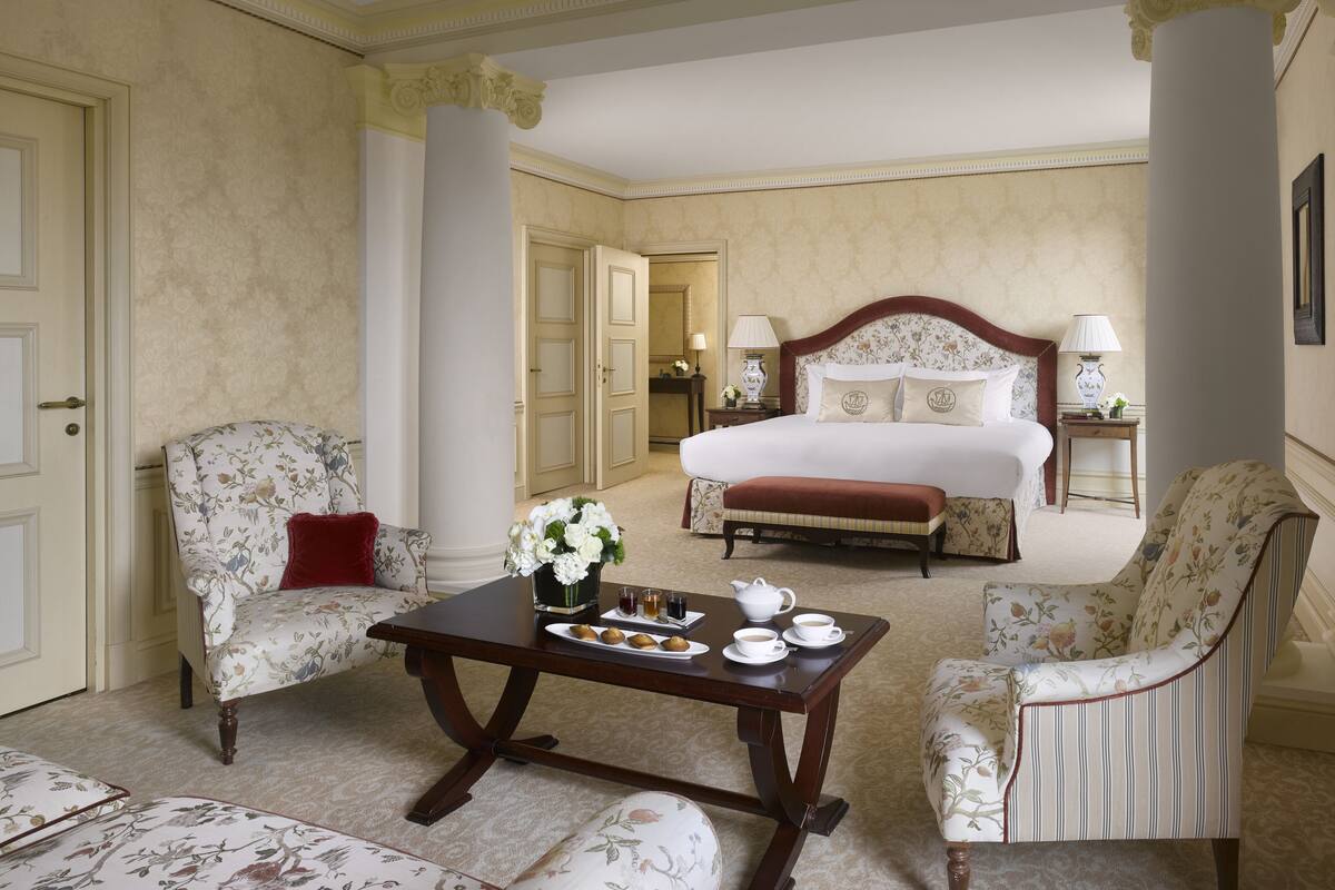 A hotel room with a large bed, two armchairs, a coffee table with pastries, and a vase of flowers.