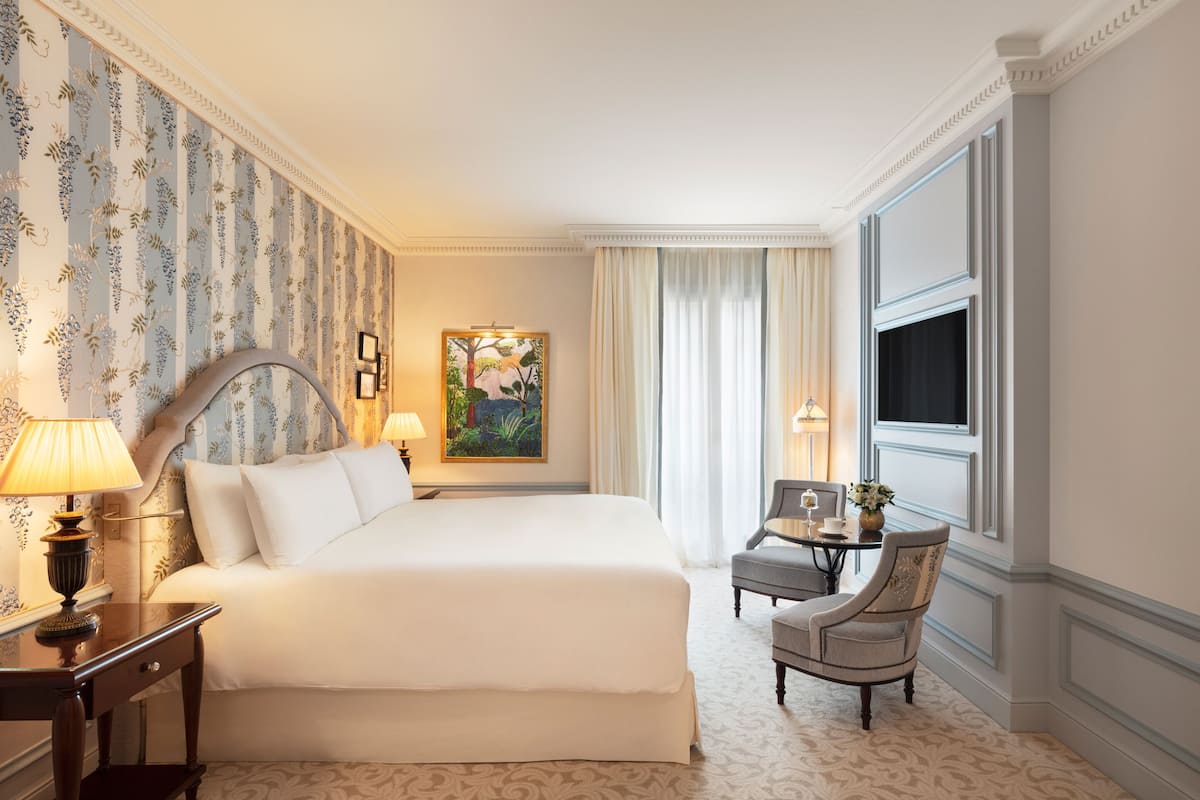 A hotel room with a large bed, a desk with a chair, a painting on the wall, and a window with curtains.