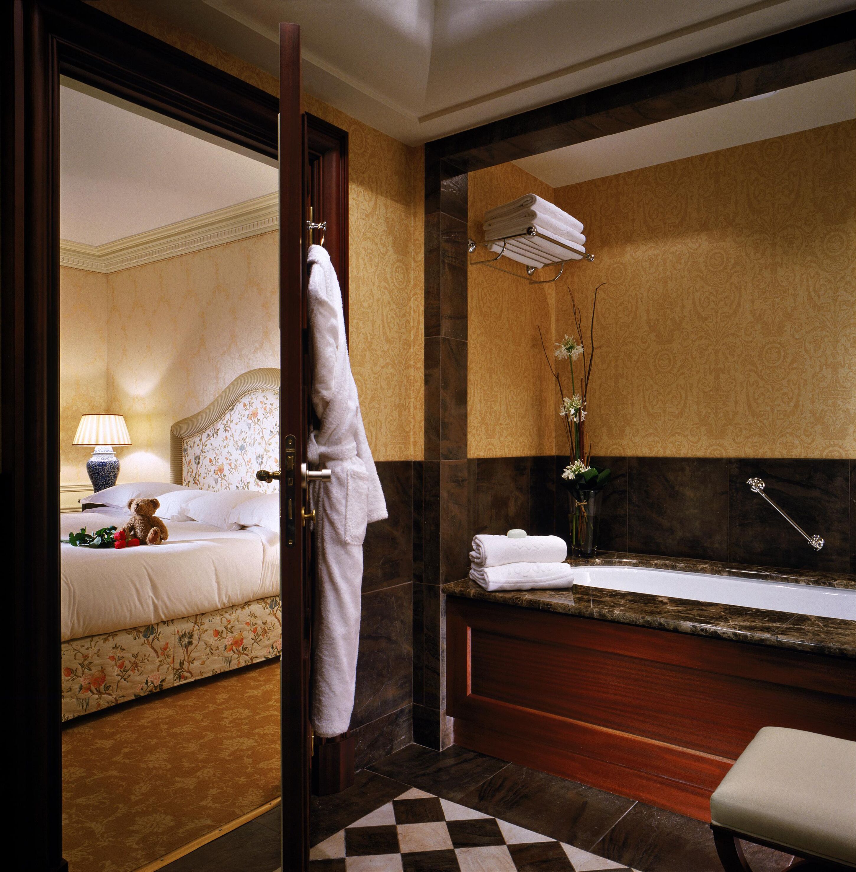 A hotel room with a bed, a nightstand, a lamp, a bathroom with a bathtub, a mirror, and a towel rack.