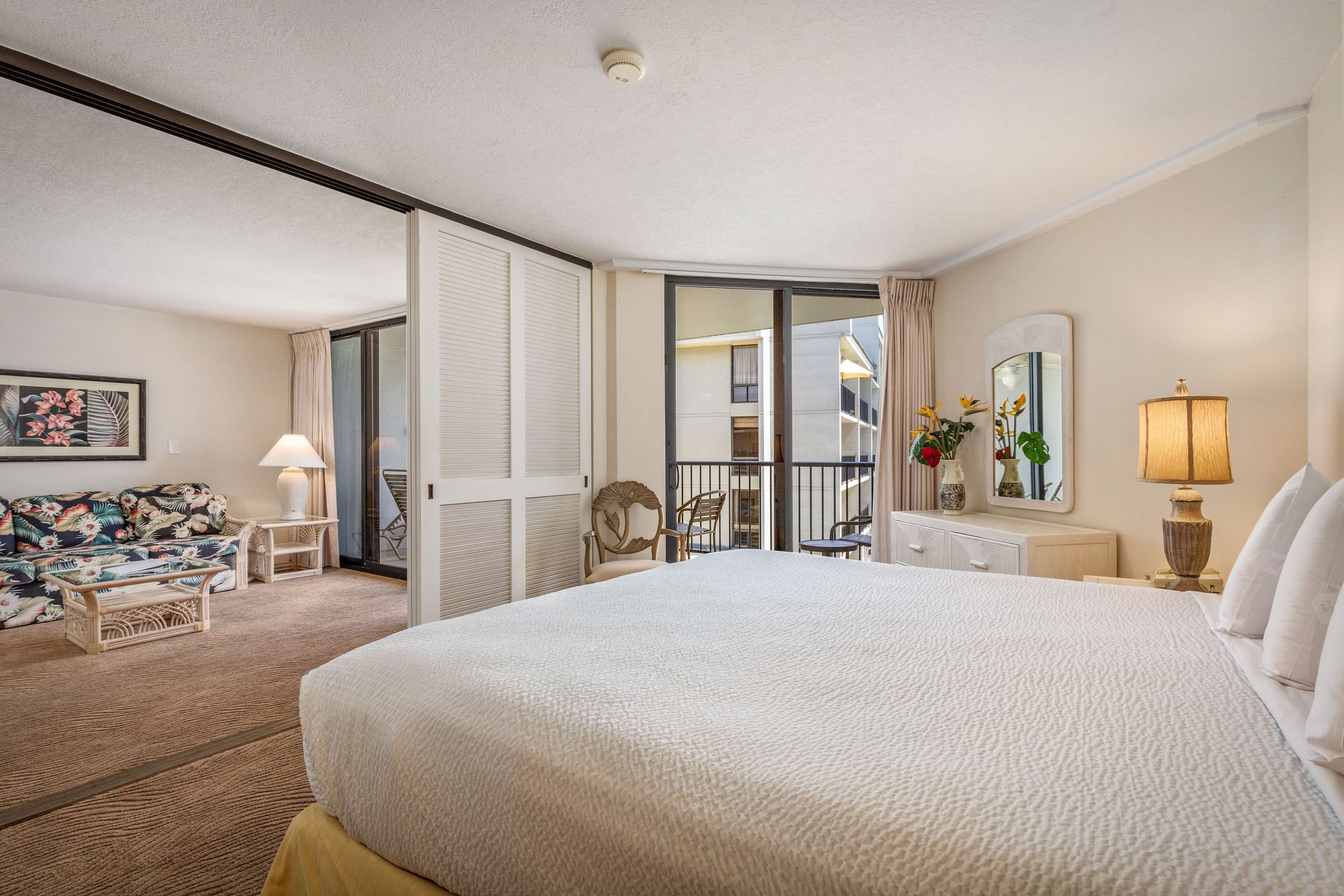 A hotel room with a large bed, a sofa, a chair, a lamp, and a view of the balcony.