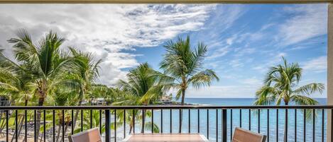 A balcony with a wooden table and chairs, overlooking a beach with palm trees and the ocean.