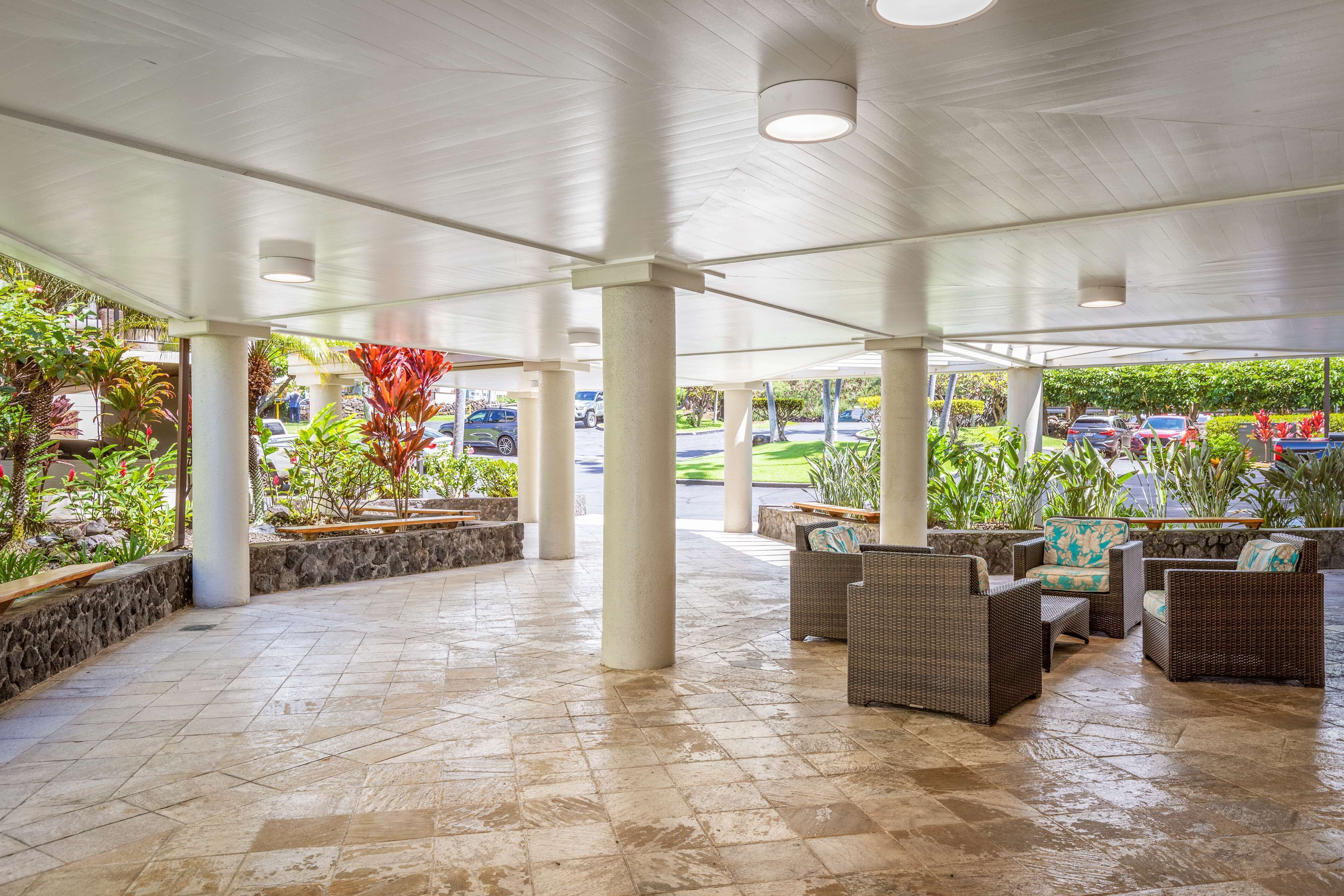 A covered outdoor area with stone flooring, white pillars, wicker furniture, and potted plants.