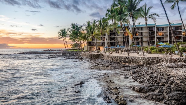 Un hôtel situé en bord de mer, doté d’un mur de soutènement en pierre, de palmiers et offrant une vue sur l’océan au coucher du soleil.