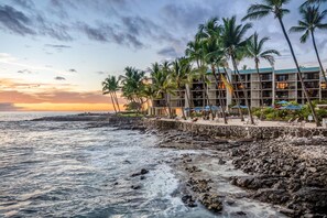 Un hôtel situé en bord de mer, doté d’un mur de soutènement en pierre, de palmiers et offrant une vue sur l’océan au coucher du soleil.