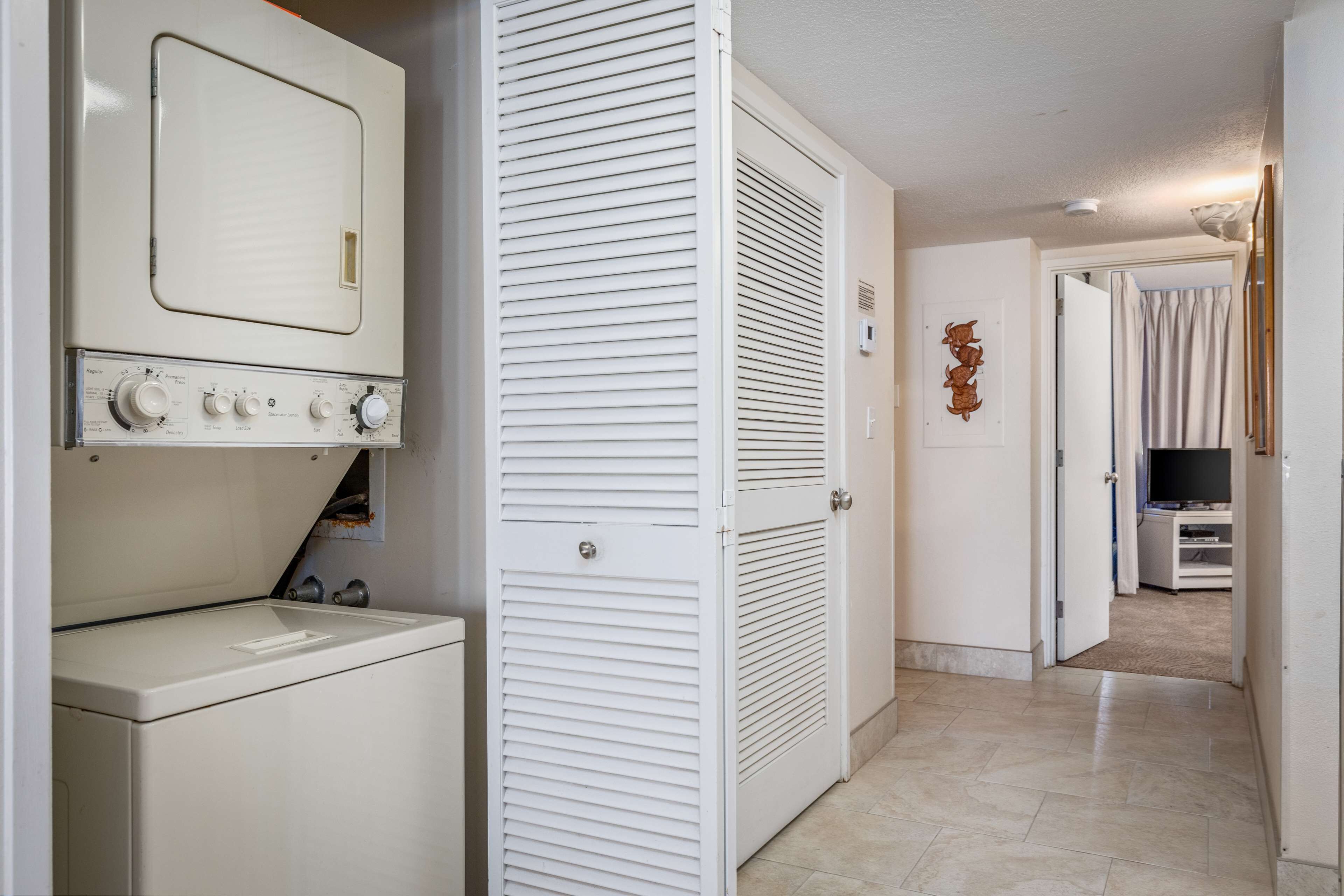 Un cuarto de lavado con lavadora y secadora, una puerta blanca con celosías y un pasillo que conduce a una sala de estar con televisor.