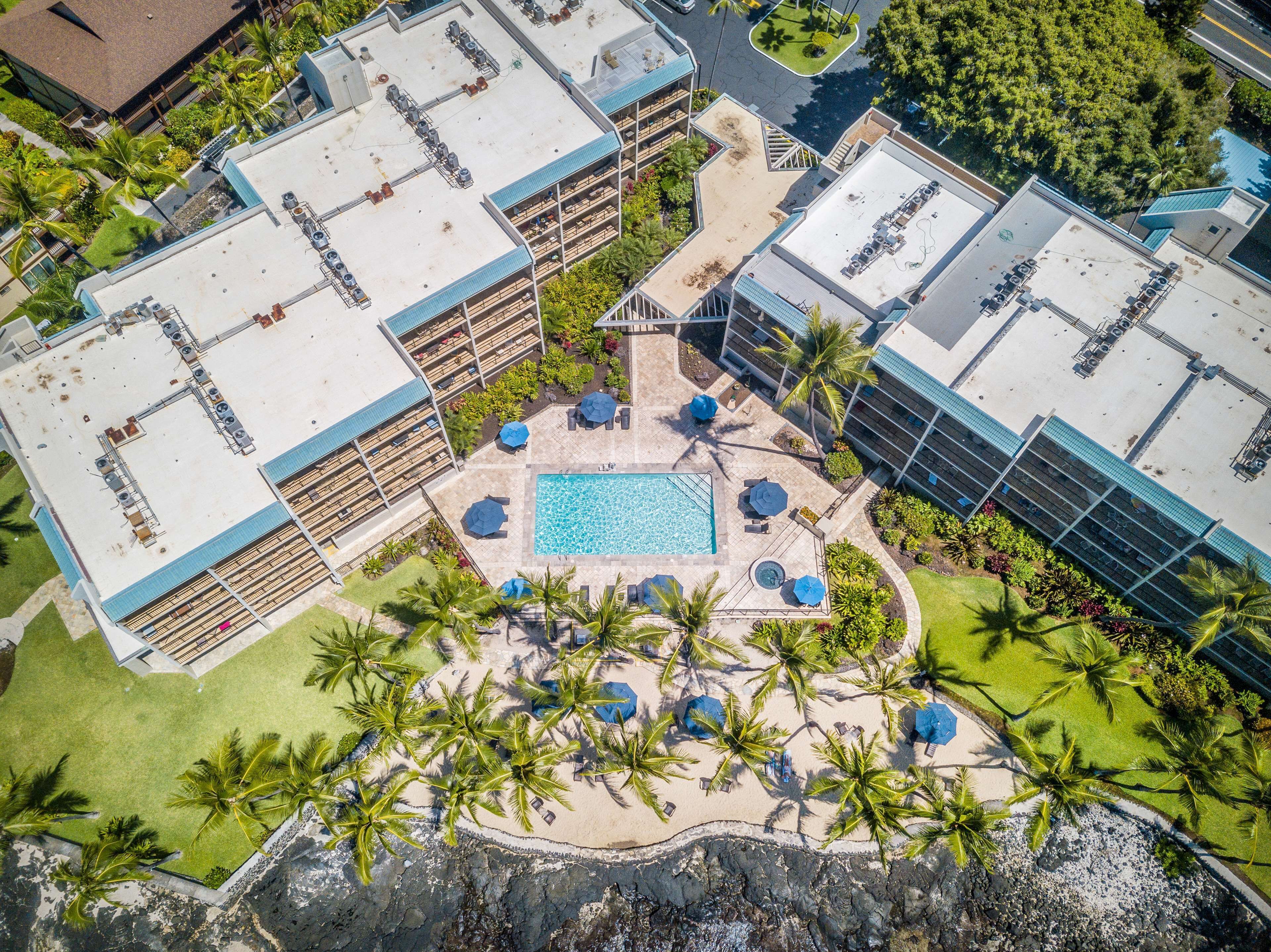 Vista aérea de un resort con varias edificaciones, un área de piscina central con tumbonas y palmeras circundantes.