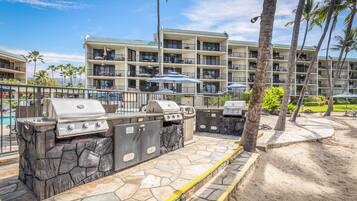 Un espace barbecue extérieur doté de plans de travail et de grils en pierre, entouré d’une piscine et d’un bâtiment d’hôtel avec des balcons.