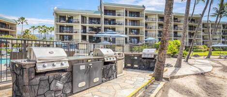 An outdoor barbecue area with stone countertops and grills, surrounded by a pool and a hotel building with balconies.