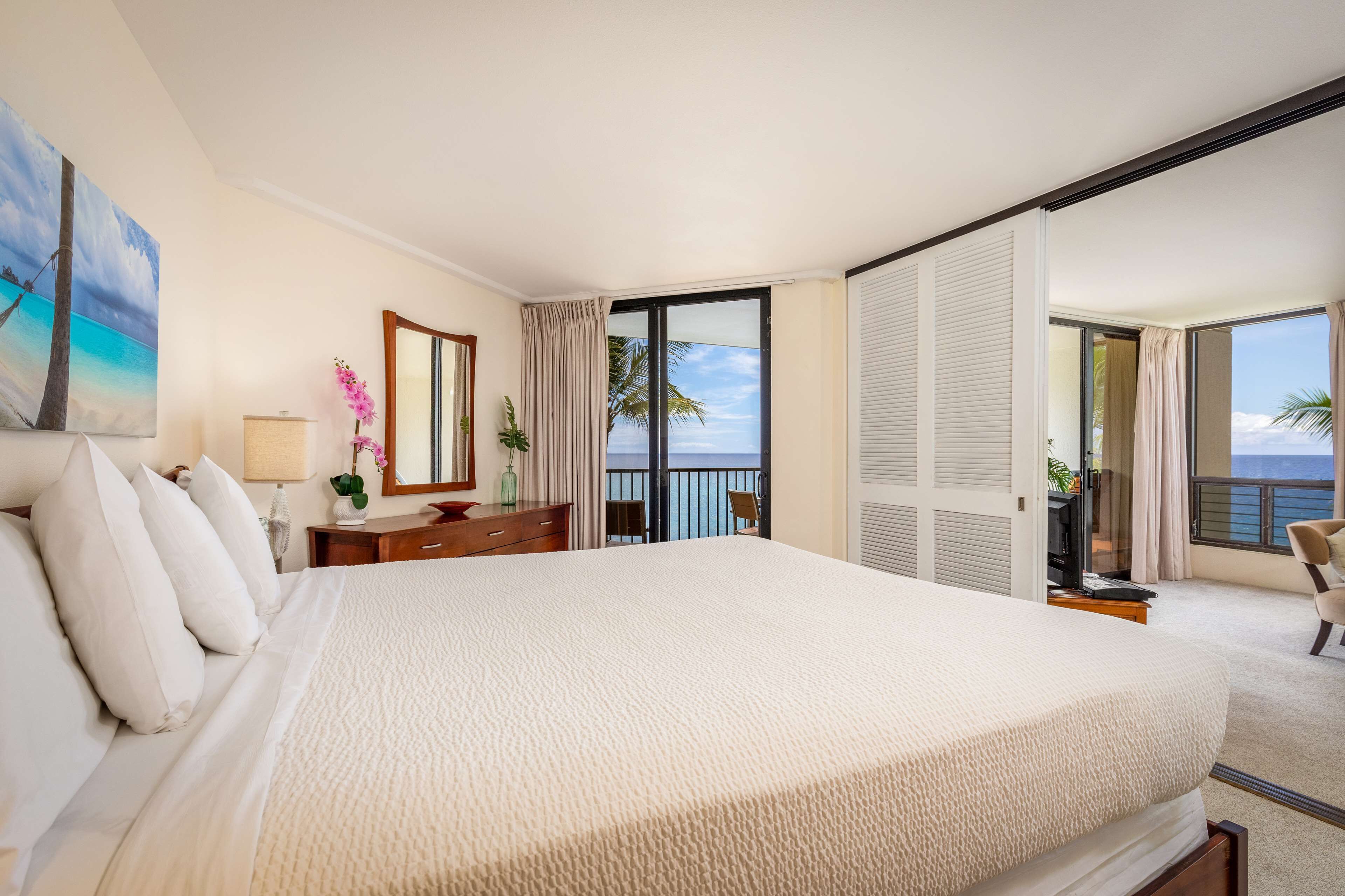 A hotel room with a large bed, a desk, a chair, a TV, and a balcony with a view of the ocean.