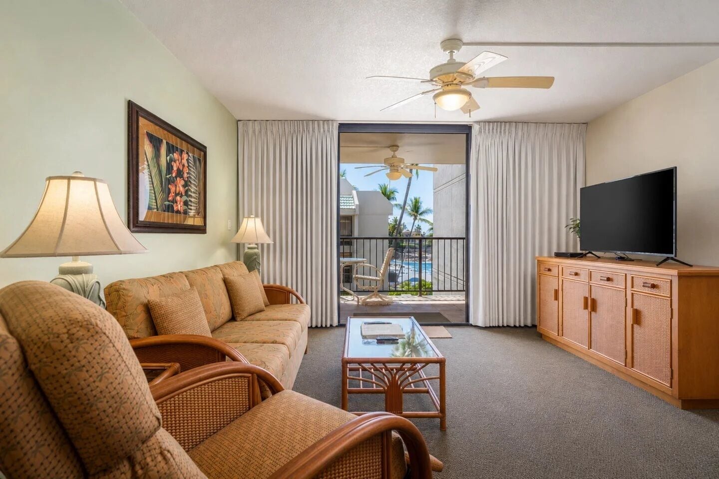 A hotel room with a sofa, chairs, a television, a ceiling fan, and a view of the ocean through the balcony.