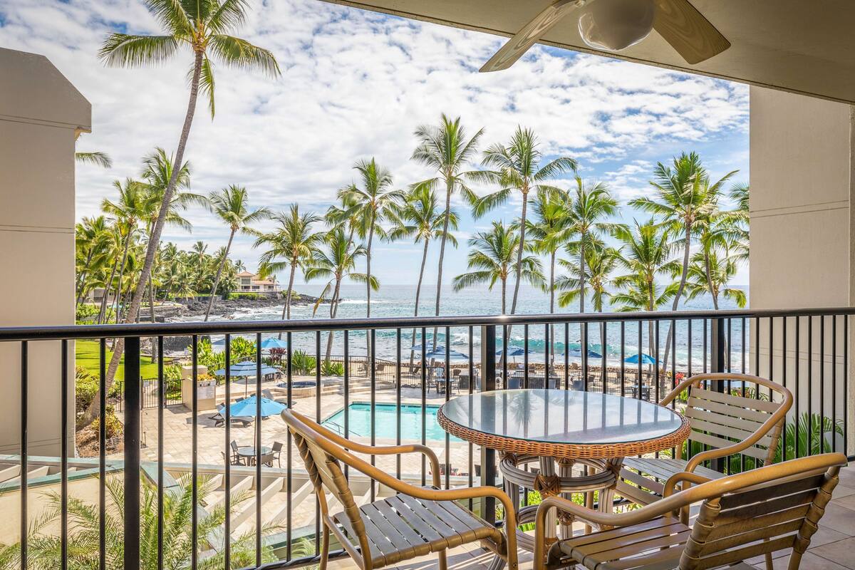 Un balcón con vista a una playa y palmeras, una piscina y mobiliario exterior.