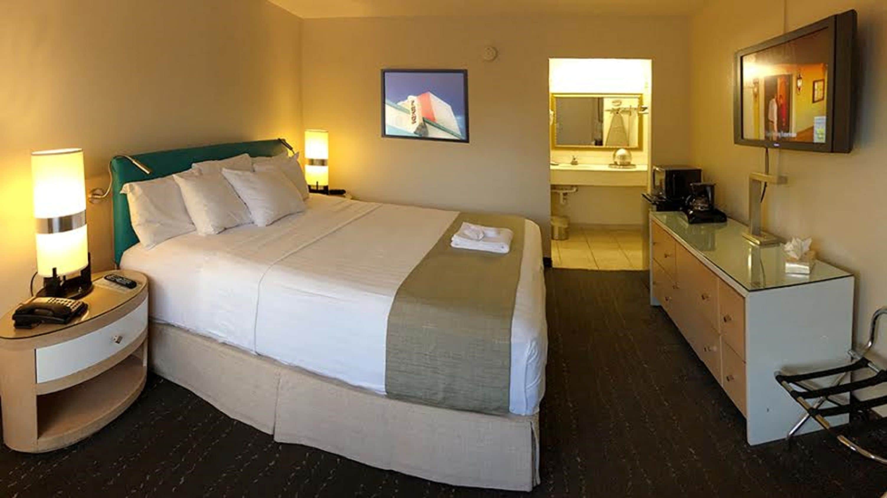 Une chambre d’hôtel avec un lit, des tables de chevet, une commode, un miroir et une salle de bain visible à travers une porte ouverte.