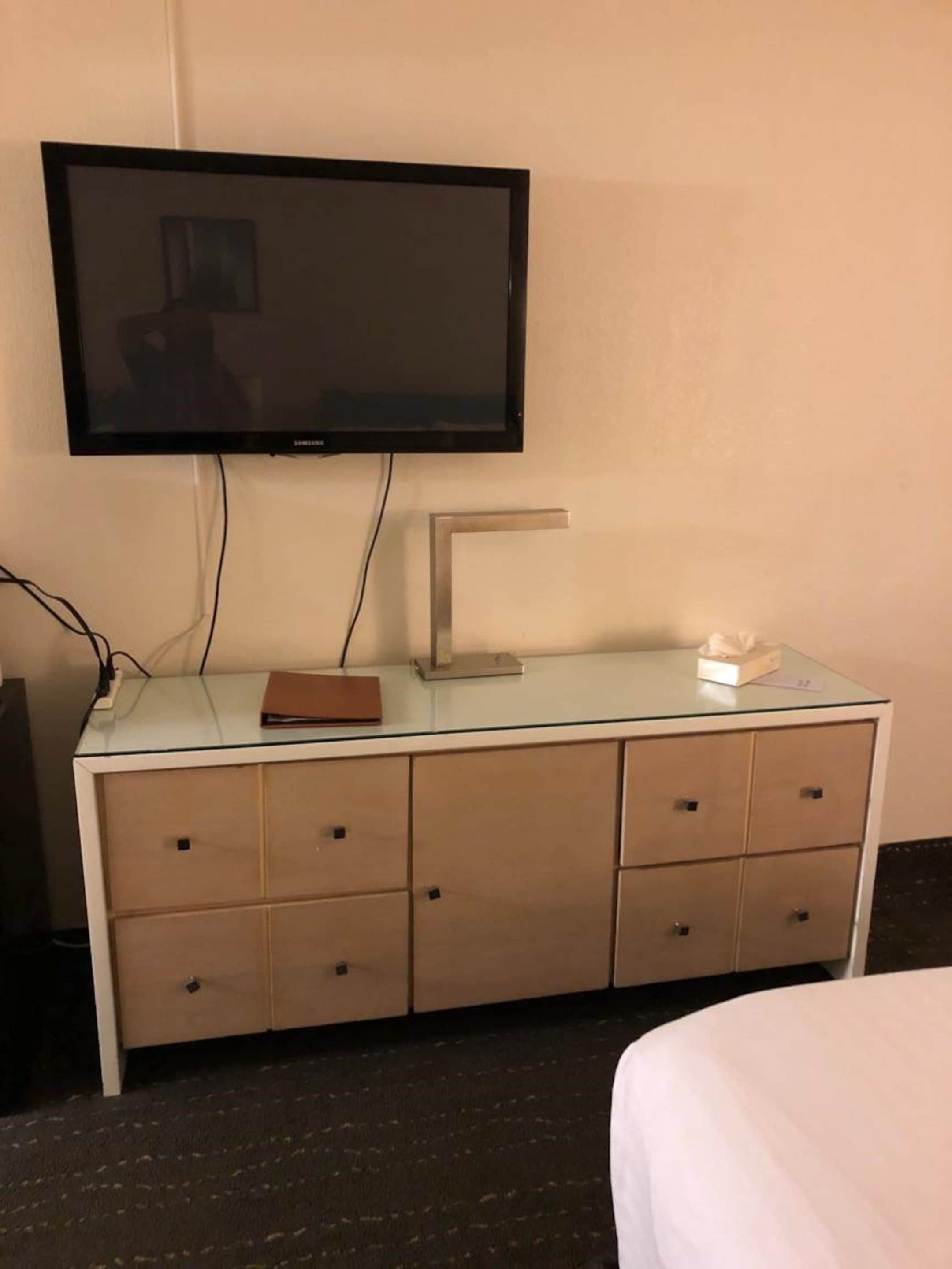 Habitación de hotel con televisor de pantalla plana montado en la pared, un escritorio de vidrio con una computadora portátil, una caja de pañuelos y una carpeta marrón.