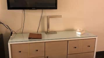 Habitación de hotel con televisor de pantalla plana montado en la pared, un escritorio de vidrio con una computadora portátil, una caja de pañuelos y una carpeta marrón.