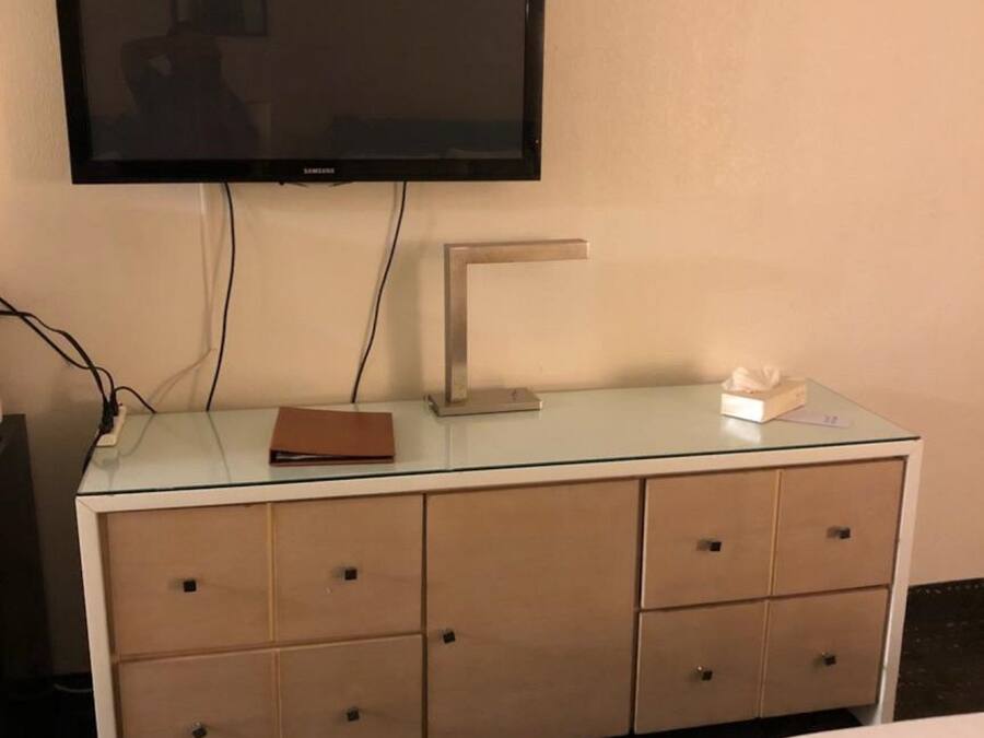 Habitación de hotel con televisor de pantalla plana montado en la pared, un escritorio de vidrio con una computadora portátil, una caja de pañuelos y una carpeta marrón.
