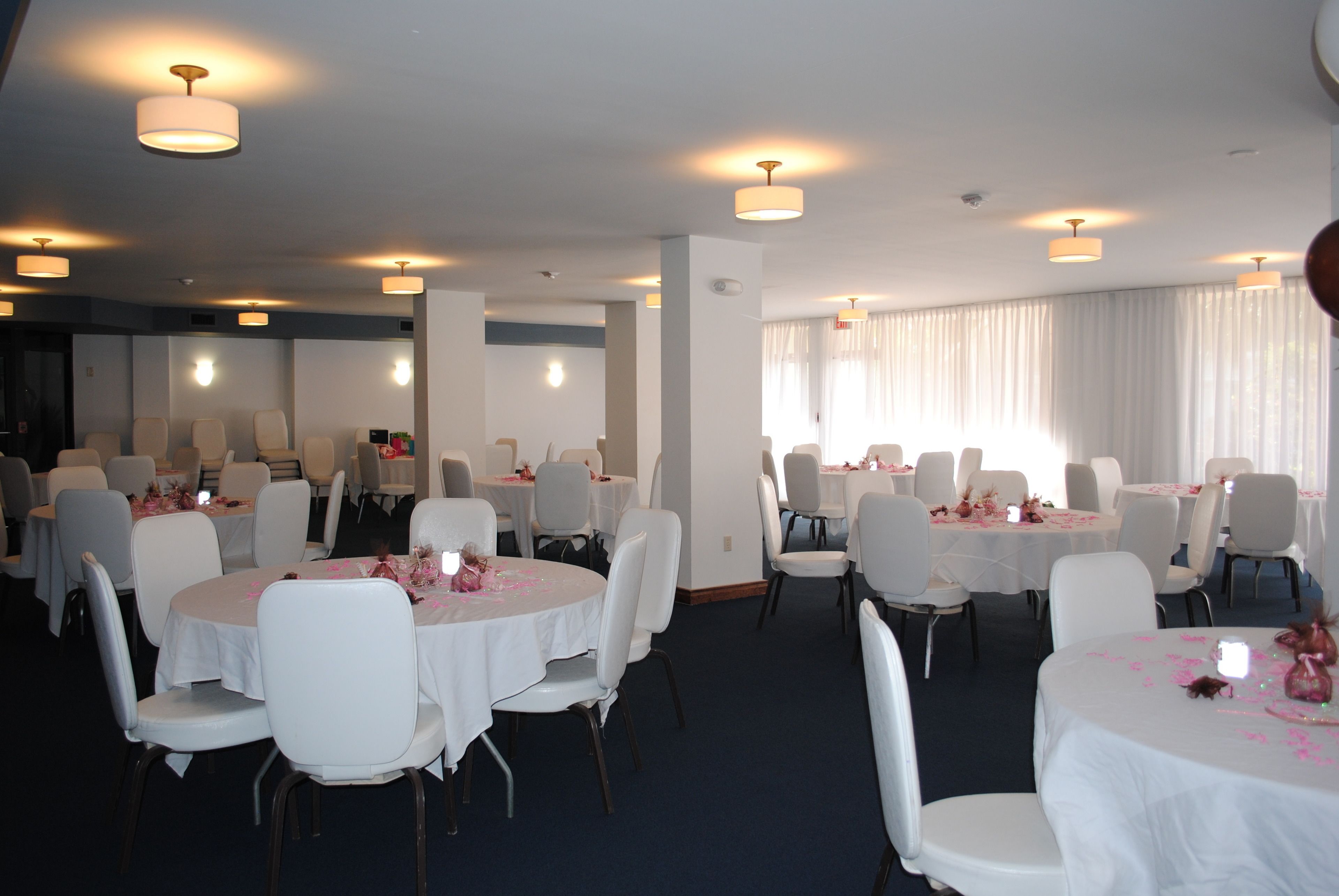 Uma sala de banquetes com mesas redondas e cadeiras brancas, com luminárias no teto e grandes janelas com cortinas.