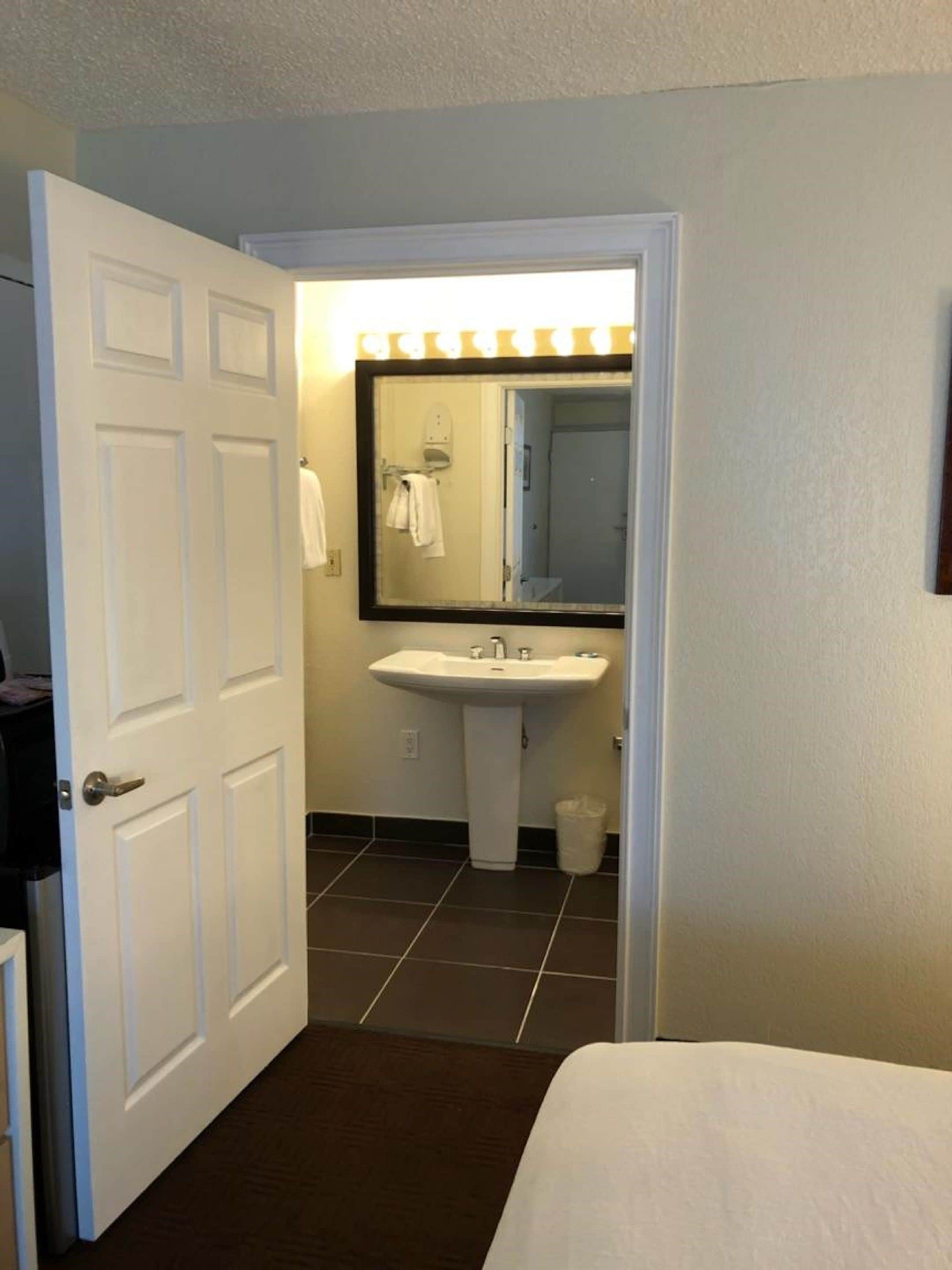 Un bagno d'albergo con un lavandino bianco, uno specchio e una porta che dà su un'altra stanza.
